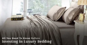 Luxury bedroom featuring silky champagne-colored bedding, matching pillowcases, a neatly styled bed, bedside table with a lamp and alarm clock, and large windows providing abundant natural light.