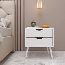 Modern white two-drawer bedside table with tapered wooden legs, styled with a small decorative vase and bowl beside an upholstered bed in a minimalist bedroom.