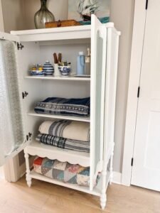 White freestanding storage cabinet with open doors displaying neatly folded blankets, towels, decorative bowls, and bedroom essentials on organized shelves.