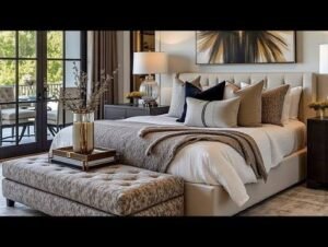 Luxurious modern bedroom featuring a beige upholstered bed with layered neutral bedding, a tufted bench, elegant bedside lamps, framed wall art, and large glass doors that fill the room with natural light.