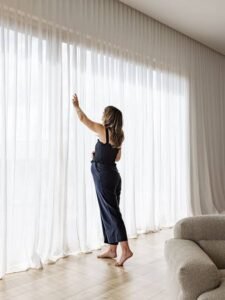 Woman adjusting sheer white floor-to-ceiling curtains in a bright modern living room with natural light, minimalist décor, and a curved upholstered sofa.