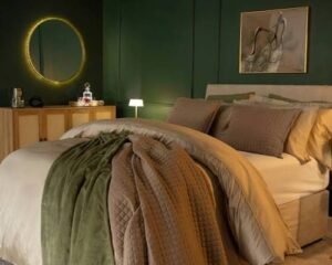 Elegant bedroom with deep green walls, layered neutral bedding, textured throws, a round backlit mirror, wooden sideboard, and soft ambient lighting creating a luxurious atmosphere.