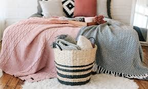 Bedroom bed styled with layered throw blankets in soft pink and gray, with additional blankets stored in a woven basket.