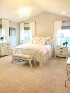 Elegant traditional bedroom with white furniture, a carved bed frame, crystal chandelier, matching nightstands, soft neutral walls, and large windows with white curtains.