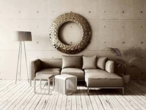 A modern minimalist living room featuring a beige sectional sofa, a geometric circular wall mirror, a floor lamp, nesting tables, and a light wood floor against a textured concrete wall.