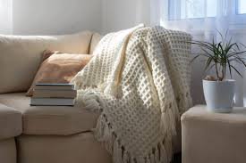 Cream knitted throw blanket casually draped over a beige sofa beside a window with books and a potted plant.