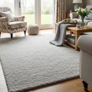 Cozy living room with a soft neutral area rug, floral armchair, wooden coffee table, knitted throw blanket, and large glass doors letting in natural light.