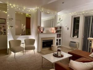Cozy modern living room with warm string lights, a white fireplace and mantel, two accent chairs, a white coffee table, soft carpet, built-in shelving, and large windows creating a warm, inviting atmosphere.