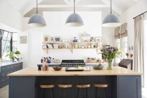 Bright modern farmhouse kitchen with a large navy blue island, wooden countertop, three gray pendant lights, open shelves, fresh flowers, and vaulted ceilings filled with natural light.