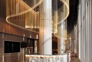 Modern luxury lobby with a marble reception desk, dramatic circular LED chandelier, vertical wood slat walls, soaring ceilings, and floor-to-ceiling windows creating an elegant contemporary atmosphere.