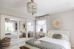 Bright contemporary bedroom with a white upholstered bed, mirrored sliding wardrobe, crystal chandelier, soft gray knitted throw, sheer curtains, and neutral décor creating a calm and elegant atmosphere.